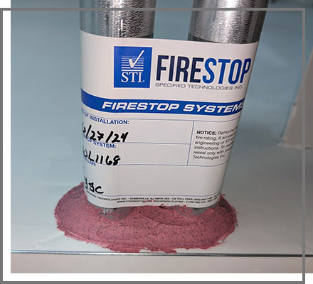Two insulated pipes pass through a floor, sealed with a pink firestop material and labeled with a tag
